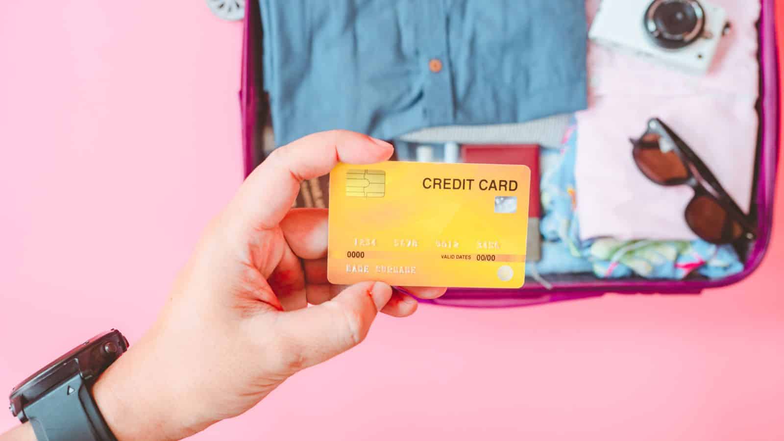 A hand holding a yellow credit card in front of an open suitcase packed with clothes, sunglasses, and a camera, with a pink background.