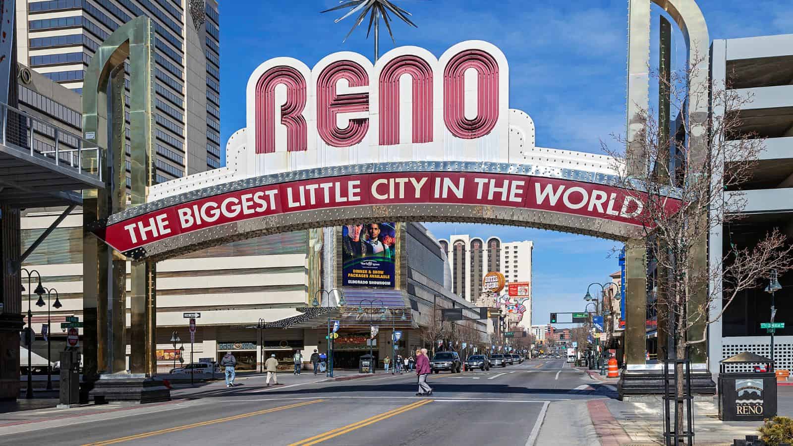 A large archway sign reads “RENO, The Biggest Little City in the World” over a downtown street lined with buildings under a clear blue sky. Pedestrians cross the street below the sign.