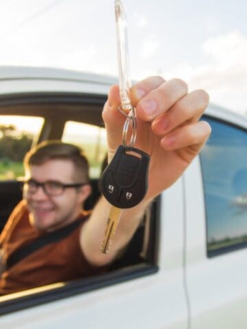A person sitting in the driver&rsquo;s seat of a white car holds a car key out of the window, smiling. The background includes greenery and a cloudy sky.