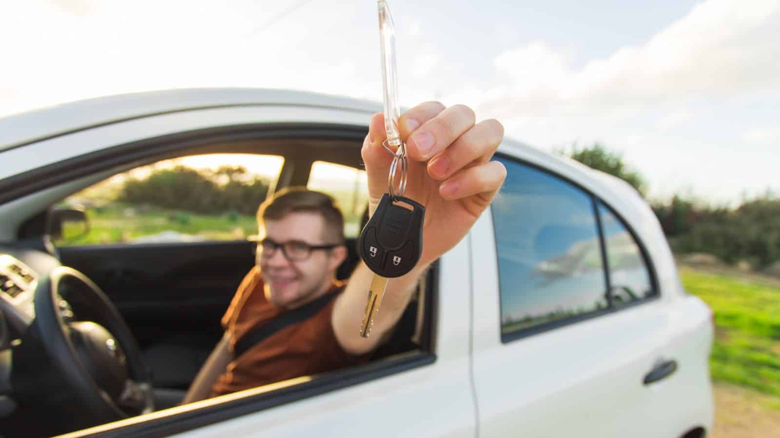 A person sitting in the driver’s seat of a white car holds a car key out of the window, smiling. The background includes greenery and a cloudy sky.