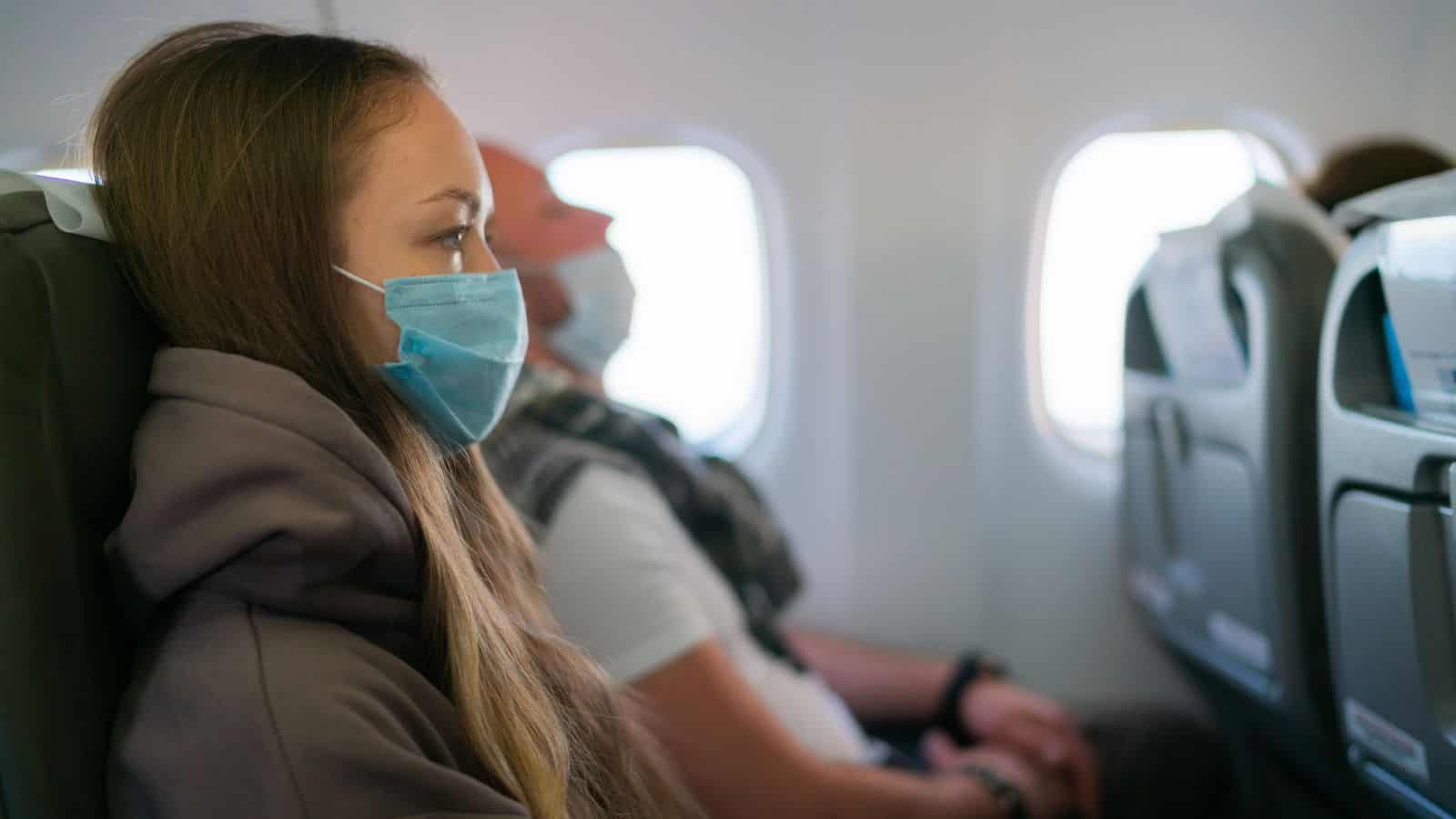 A woman and a man wearing face masks are seated side by side on an airplane. The woman is in the foreground, looking out the window, while the man sits next to her. Airplane seats and windows are visible.