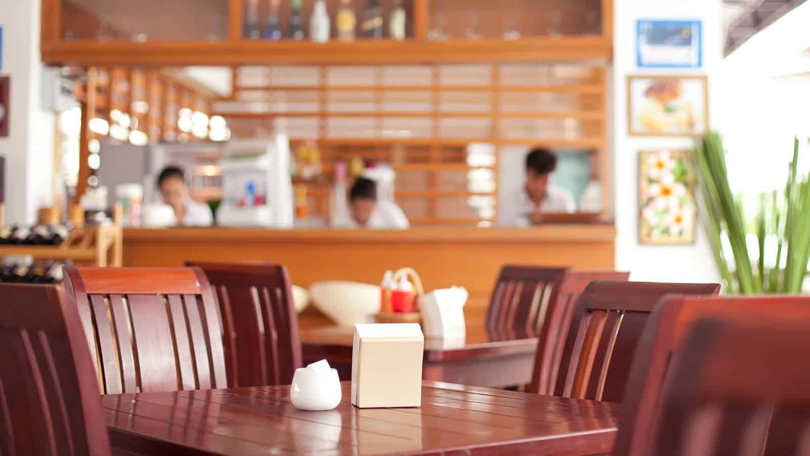 A wooden table with a white sugar bowl and a napkin holder in the foreground, with several empty wooden chairs. In the background, three people stand behind a counter in a brightly lit restaurant.