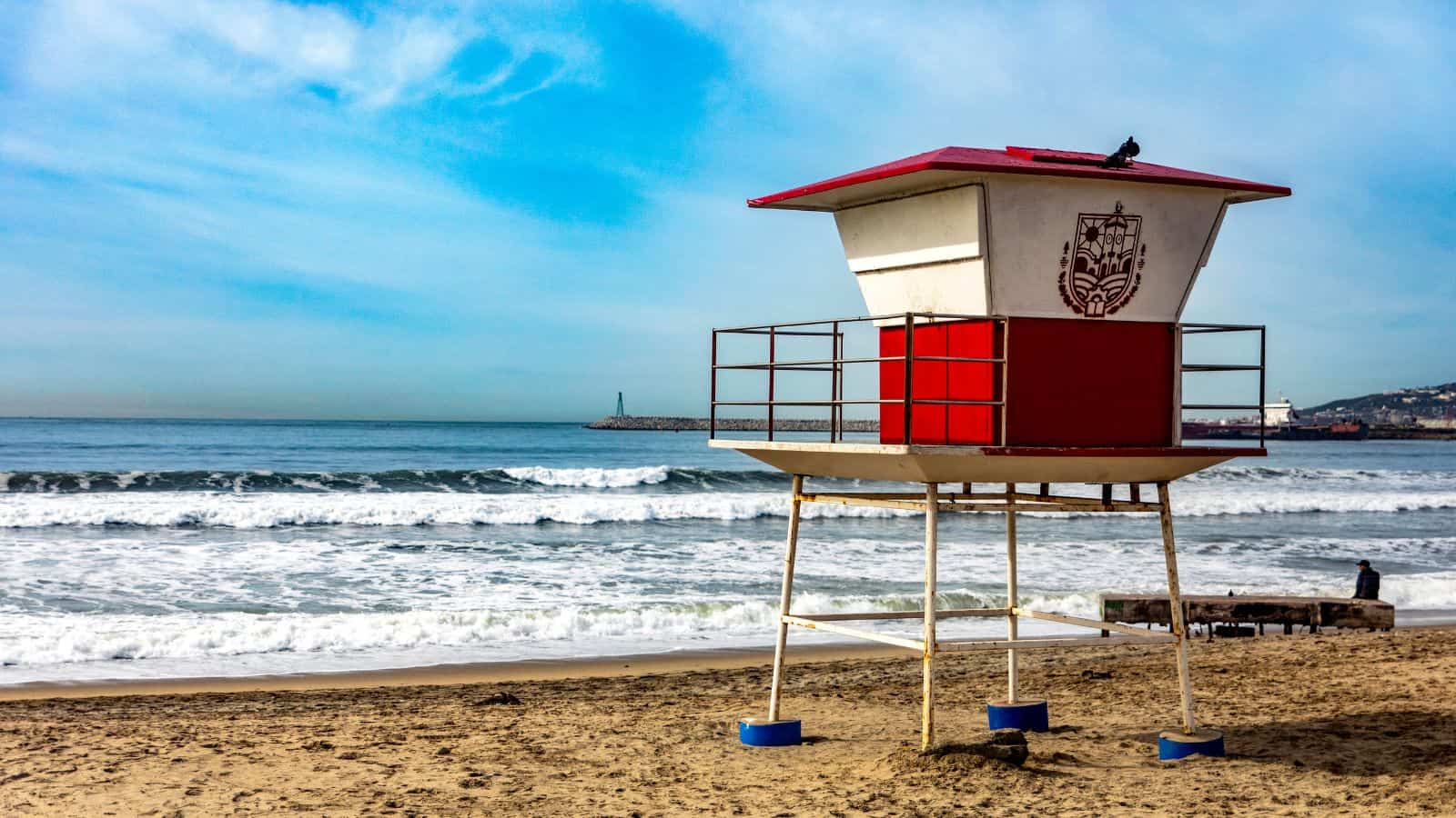A red and white lifeguard tower stands on a sandy beach near the shoreline, with waves in the background and a partly cloudy blue sky overhead. A distant pier and hills are visible on the horizon.