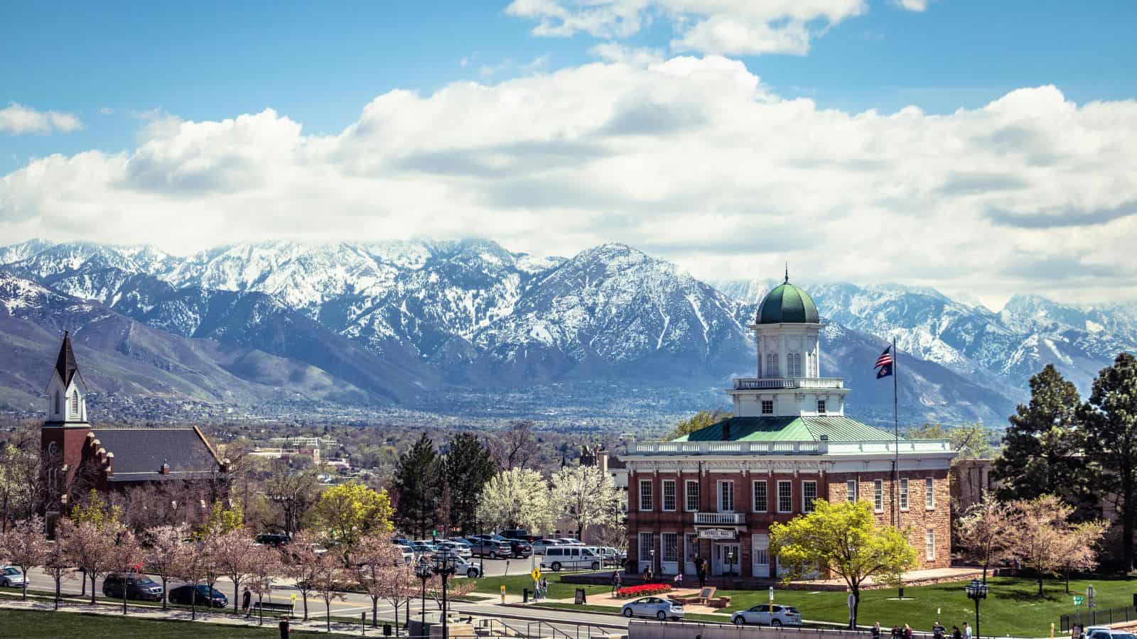 A historic brick courthouse with a green dome sits in front of snow-capped mountains under a partly cloudy sky. Trees with spring foliage and parked cars are visible in the foreground.