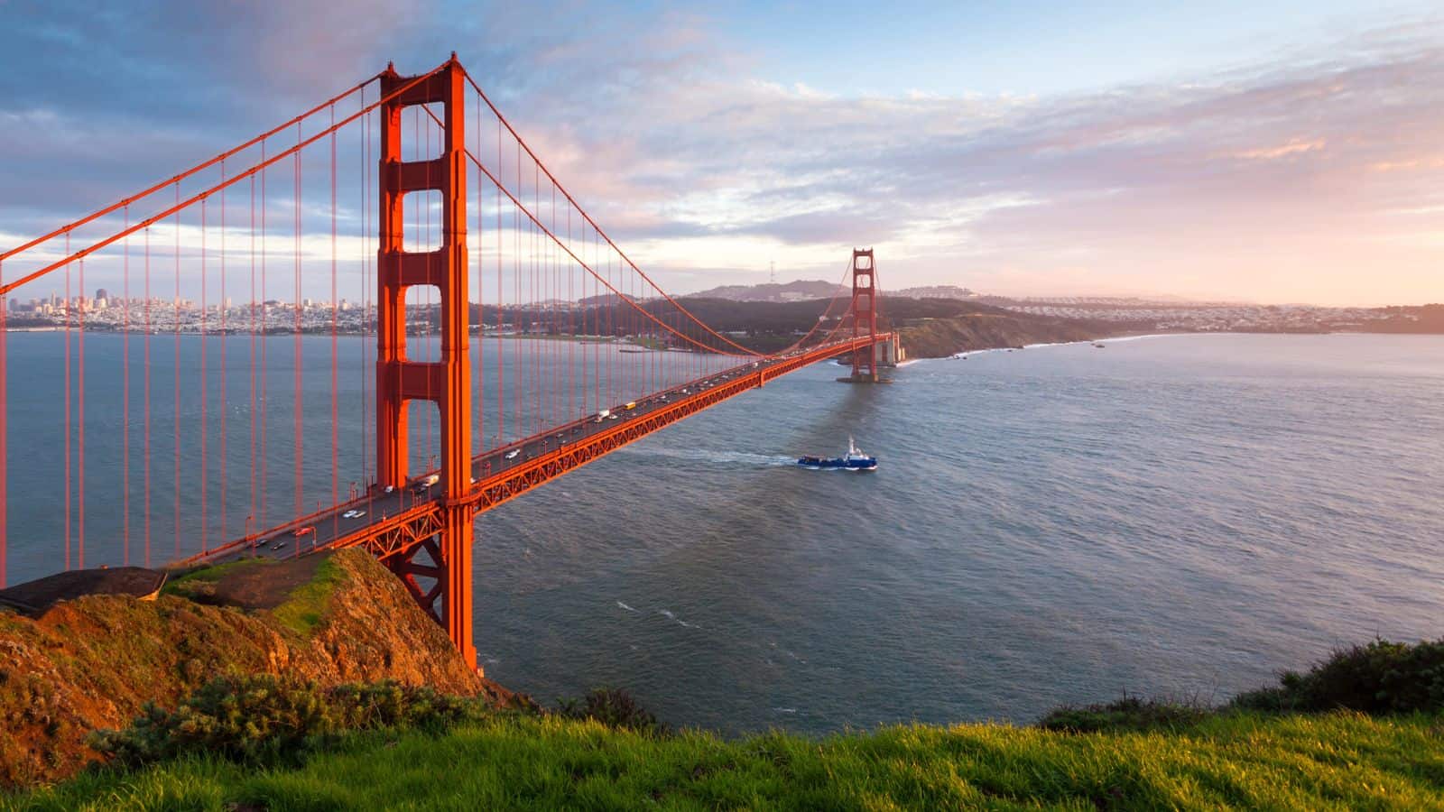 The Golden Gate Bridge spans across the water with cars driving on it. A boat moves beneath the bridge, and the scene is set during daylight with partly cloudy skies and grassy hills in the foreground.