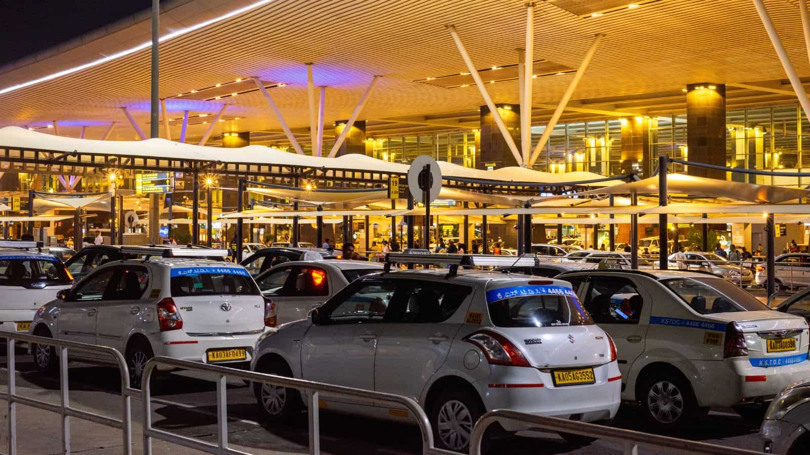 Rows of white taxis are parked outside a modern, well-lit airport terminal at night. The terminal features tall pillars, a large roof structure, and illuminated glass facades. Passengers and vehicles are visible in the background.