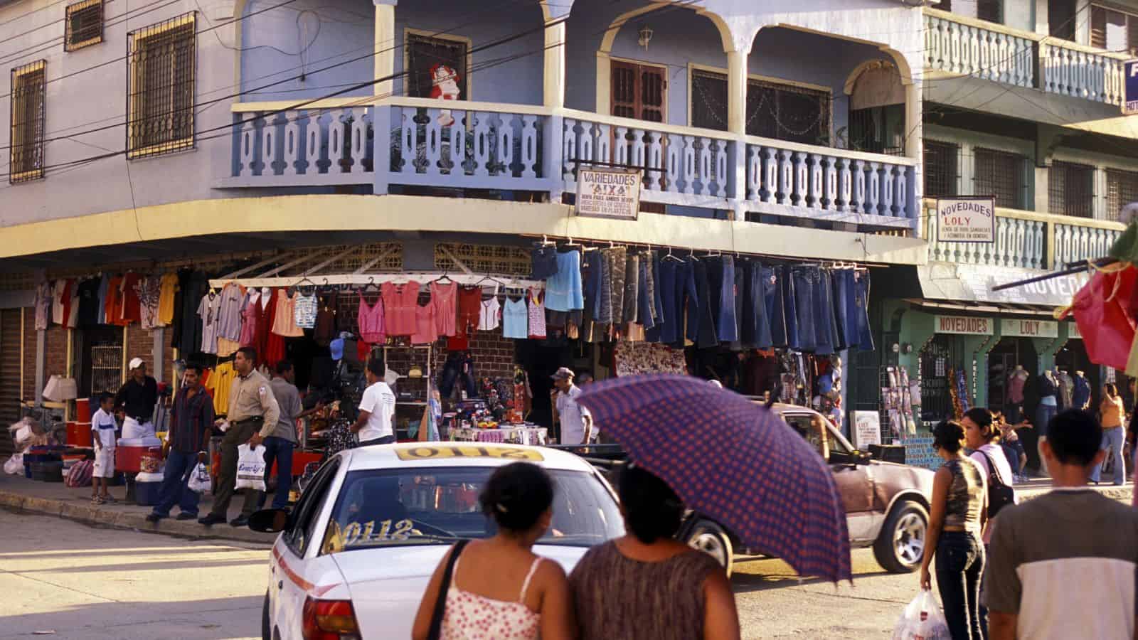 A busy street market scene shows people shopping near a building with a blue balcony. Clothing hangs on display outside stalls, and a taxi drives by while two women cross the street, one holding an umbrella.