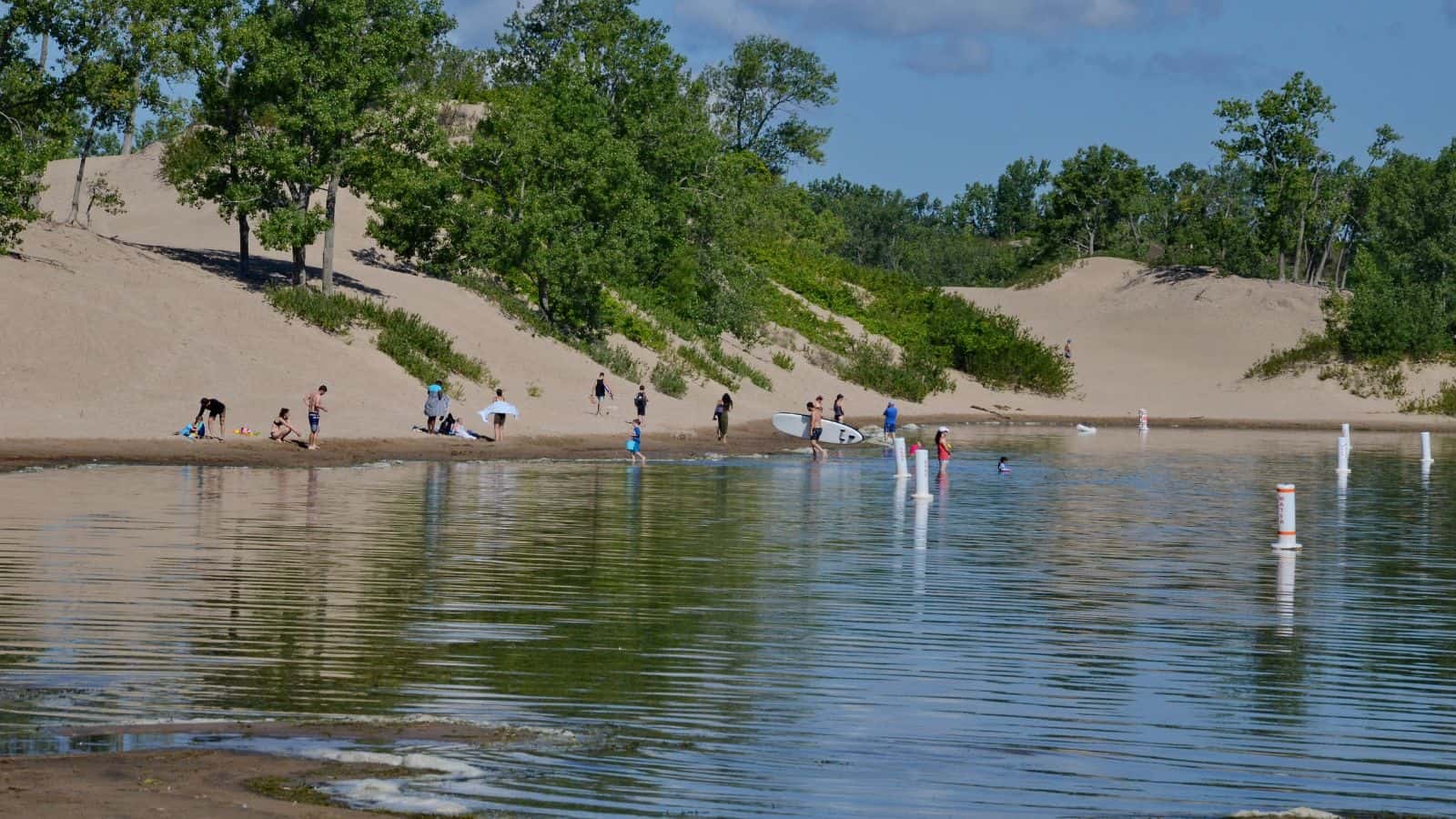 People are enjoying a sandy lakeshore with green trees in the background. Some are wading or swimming in the water, while others stand or sit on the beach. The sky is mostly clear with some clouds.