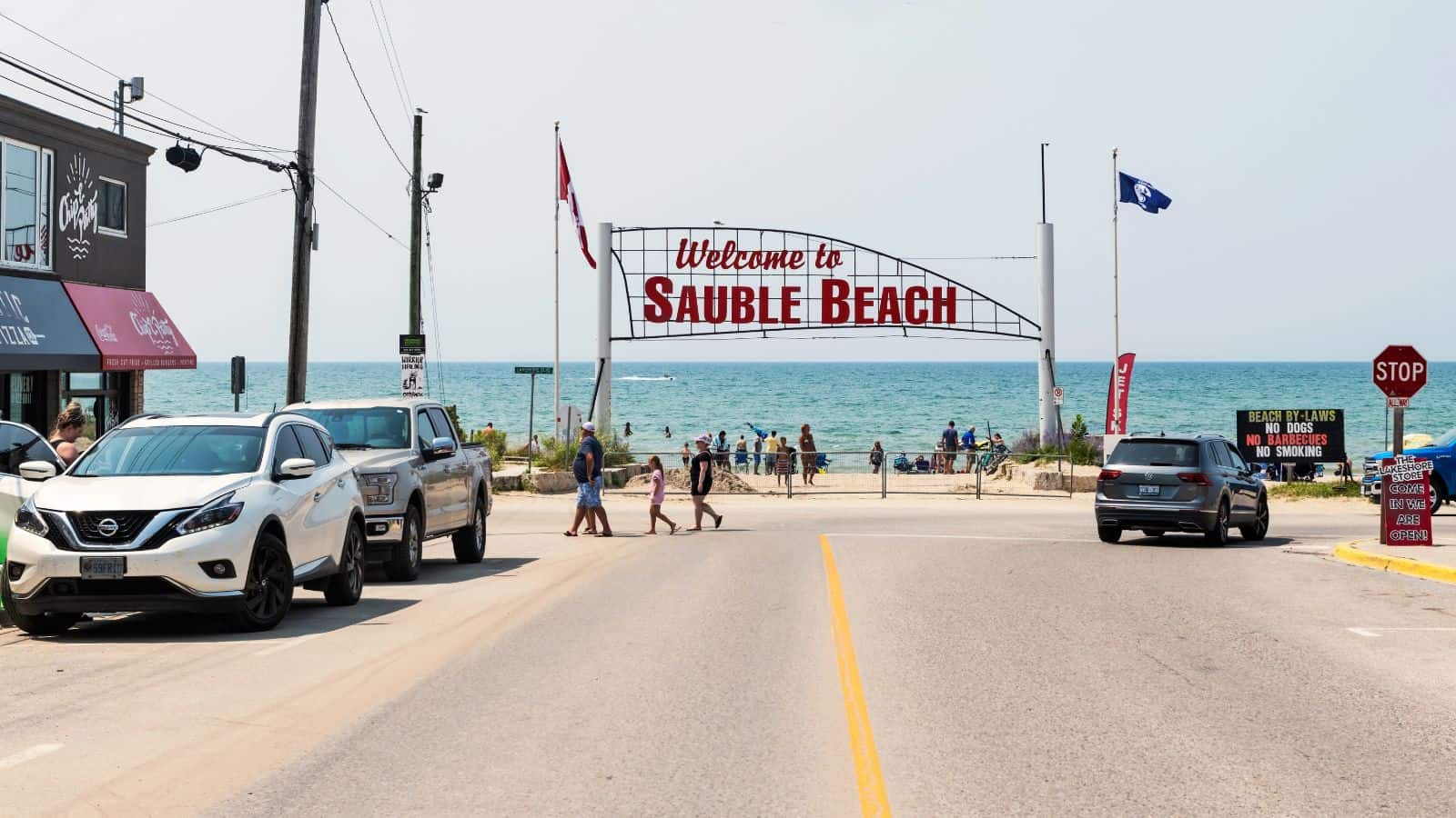 Street view of the entrance to Sauble Beach with a large "Welcome to Sauble Beach" sign, parked cars on both sides, people walking, and the lake visible in the background under a clear sky.