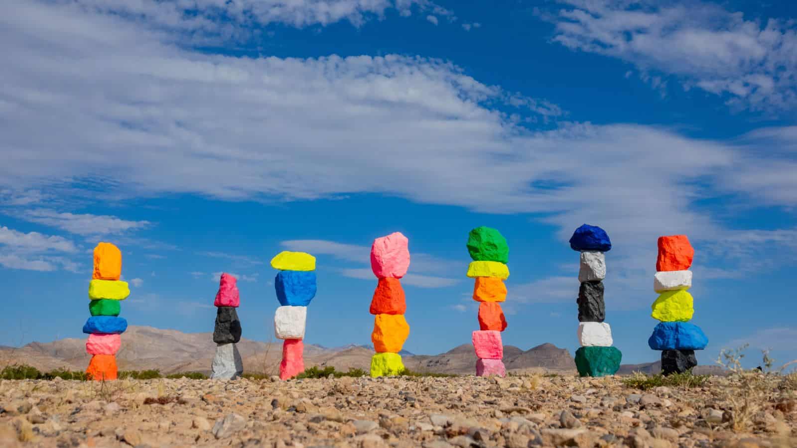 Seven vertical stacks of brightly colored, painted boulders stand in a desert landscape under a blue sky with scattered clouds and distant mountains in the background.