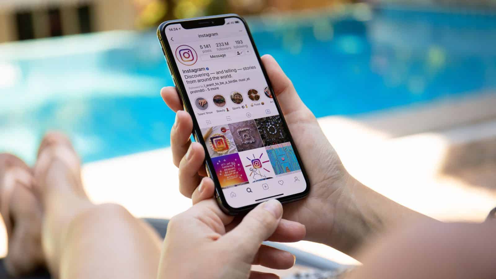 A person holds a smartphone displaying an Instagram profile while sitting beside a blue swimming pool. The screen shows profile details and several photo posts.