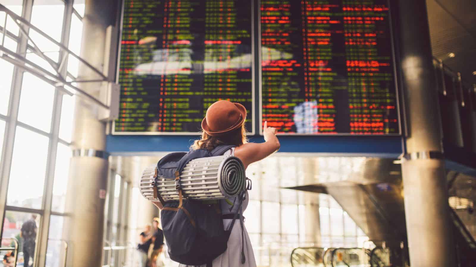 A person wearing a brown hat and backpack with a rolled sleeping mat looks up and points at a large electronic departure board displaying flight information in an airport terminal.