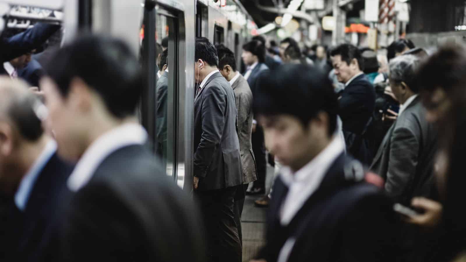 A crowded train platform with people in business attire waiting to board a train. Some individuals are looking at their phones, while others face the train doors. The scene appears busy and focused.