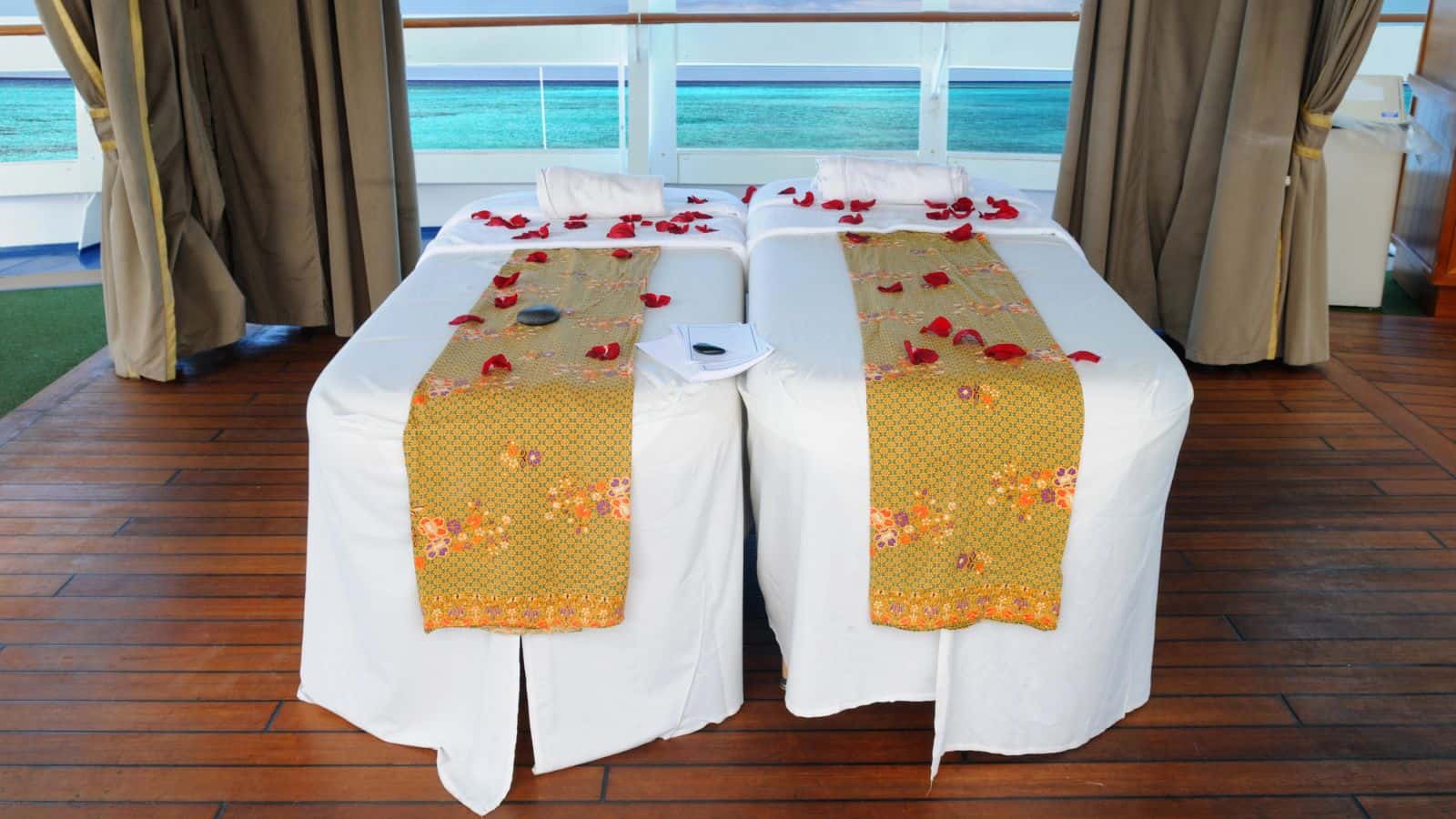 Two massage tables with yellow decorative runners and scattered rose petals are set up side by side on a wooden deck, with towels and some spa items placed on top, overlooking a view of the ocean.