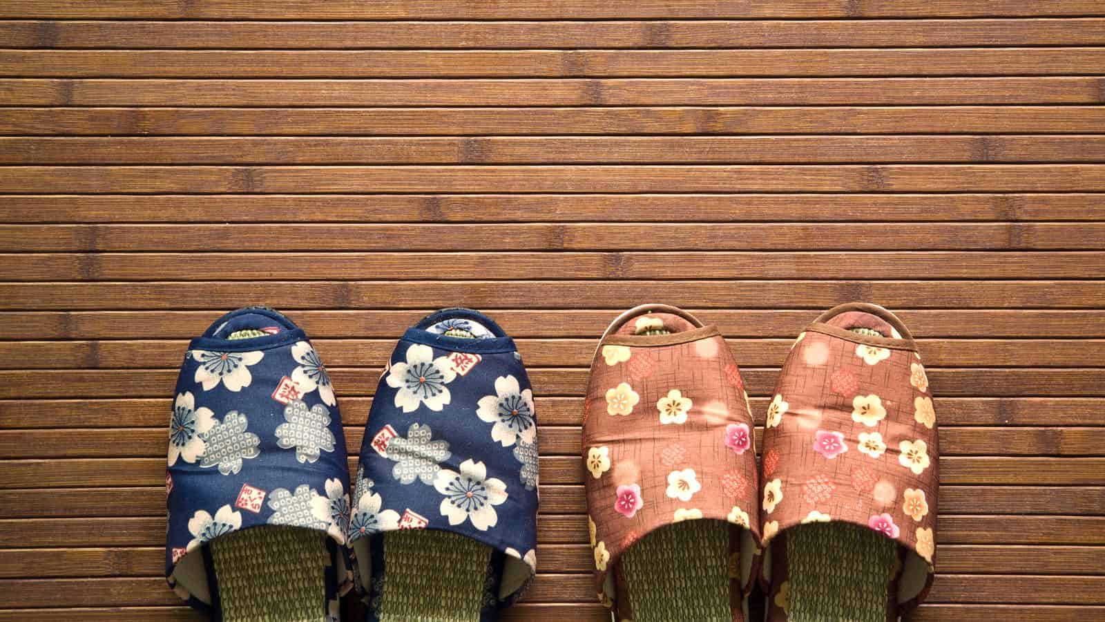 Two pairs of patterned slippers are placed side by side on a wooden floor; one pair is blue with white flowers, and the other is pink with a floral pattern.