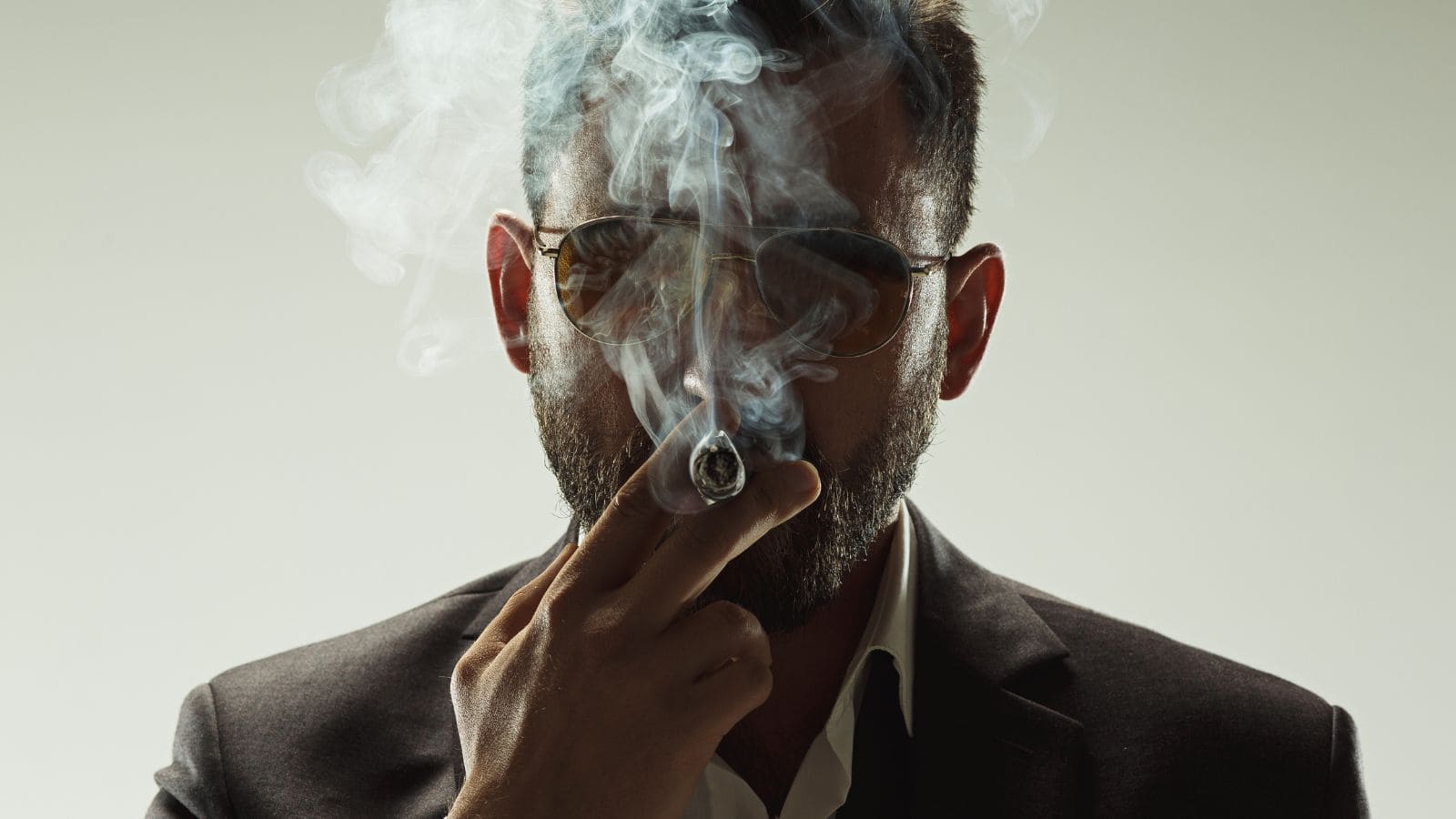 A man in a suit and sunglasses smokes a cigar, with smoke partially obscuring his face against a plain background.