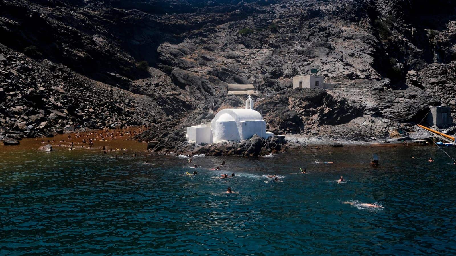 A small white chapel sits on rocky terrain at the edge of deep blue water, surrounded by swimmers. Steep, rugged cliffs rise in the background under bright sunlight.