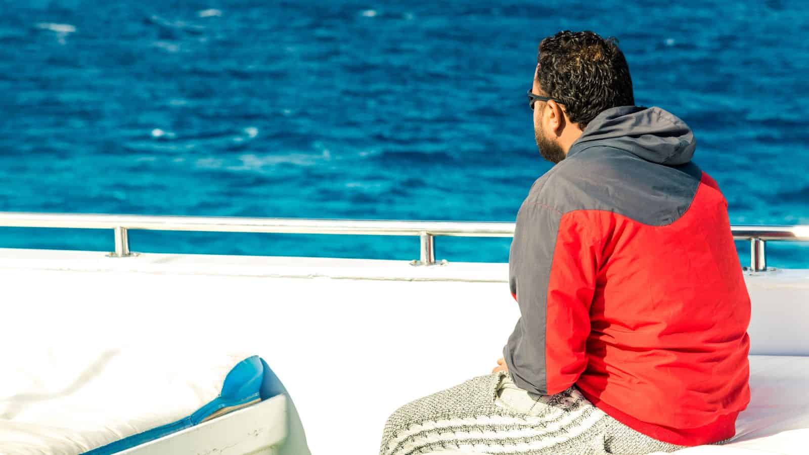 A man wearing a red and gray jacket sits on a boat, facing and looking out over the blue ocean, with sunlight illuminating the scene.