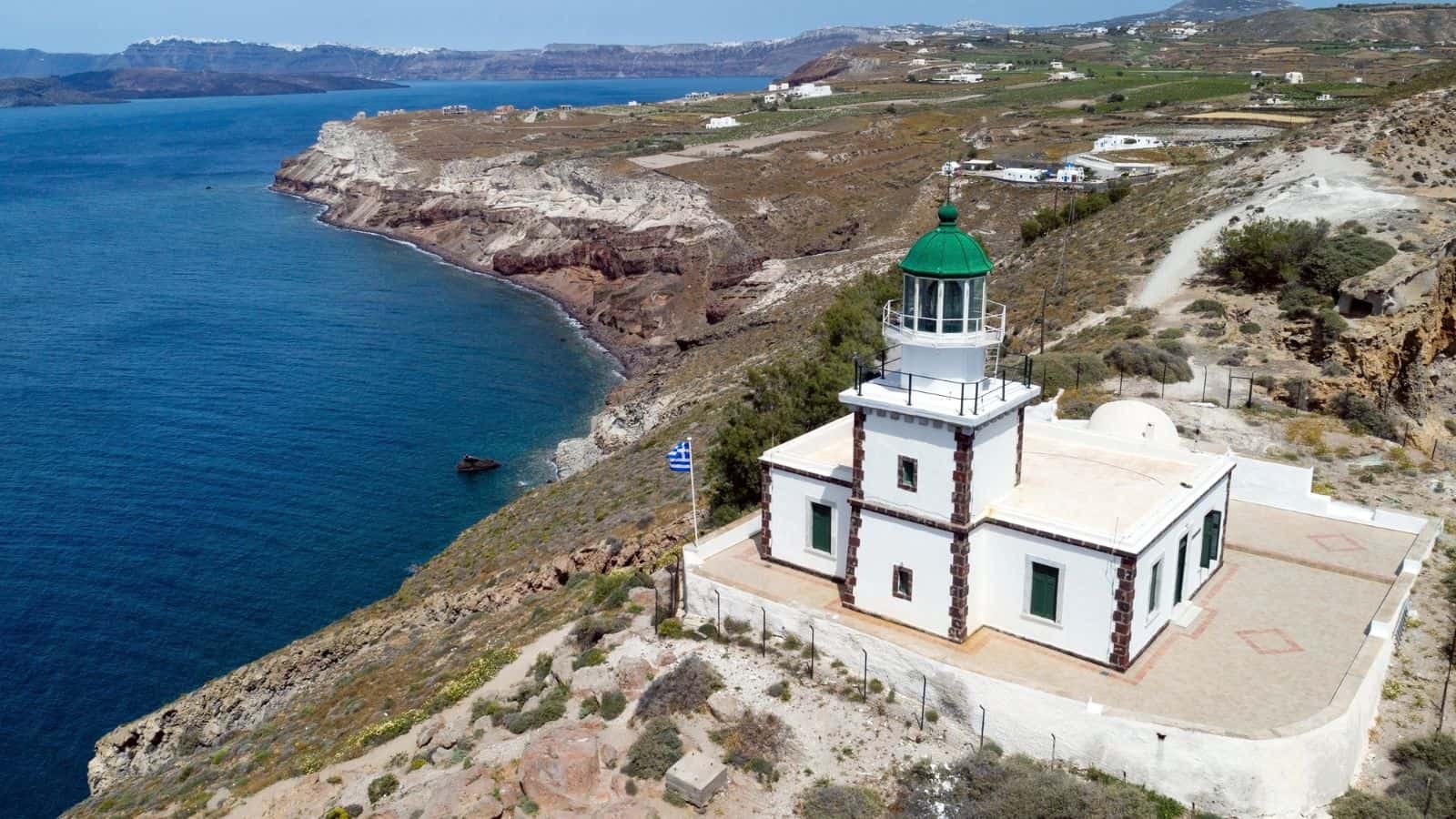 A white lighthouse with a green dome stands on a rocky coastal cliff overlooking blue water. Sparse buildings and vegetation are visible on the rugged landscape in the background. A Greek flag flies beside the lighthouse.