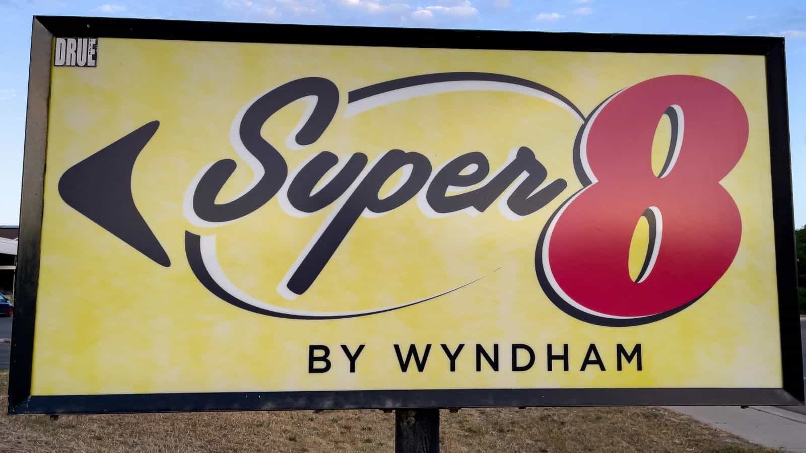 A rectangular sign with a yellow background displays the "Super 8 by Wyndham" motel logo in black, red, and yellow text. Outdoors with grass and sky behind it, this sign represents one of the US hotel chains disappointing guests.