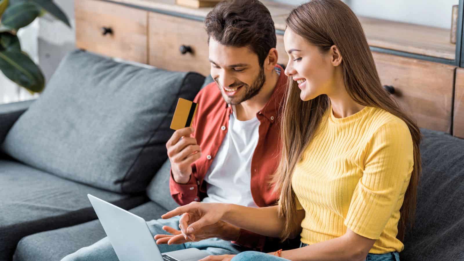 A man and woman sit on a couch with a laptop. The man holds a credit card while the woman points at the laptop screen. They appear to be making an online purchase together in a living room setting.