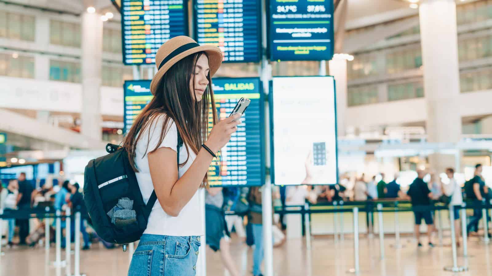A young woman with long hair, wearing a hat, backpack, and casual clothes, looks at her phone in an airport terminal. Flight information boards and people are visible in the background.
