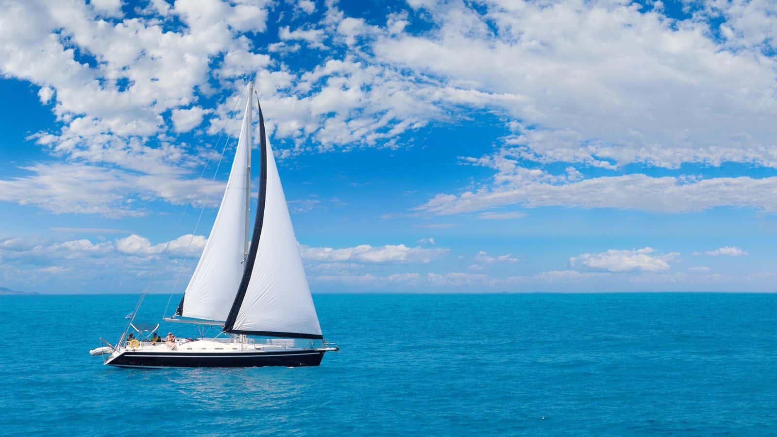 A white sailboat with tall sails moves across calm blue ocean water under a partly cloudy sky. Several people are visible on the deck, and land can be seen faintly on the horizon.