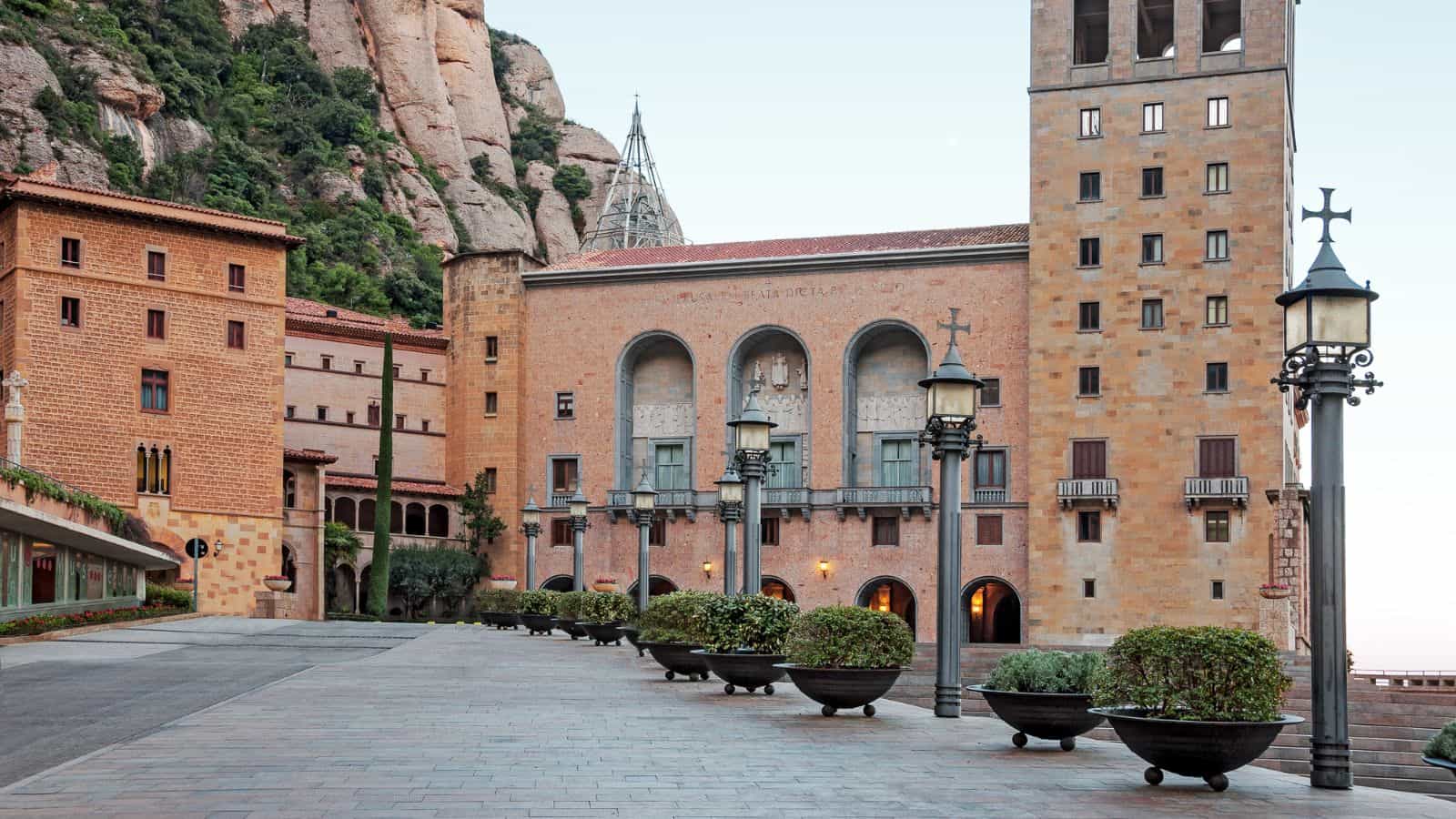 Large stone buildings with arched windows and ornate details stand at the base of a rocky mountain, with decorative street lamps and potted plants lining a paved open plaza in the foreground.