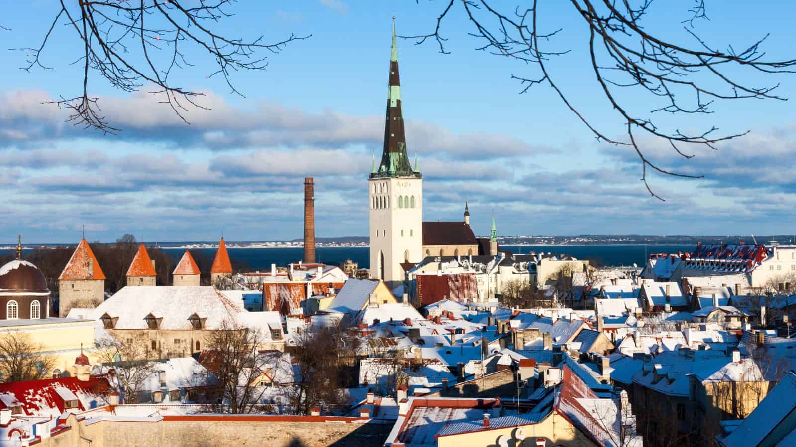 A winter view of a historic European city with snow-covered rooftops, a tall church spire, brick chimneys, and a distant body of water under a blue sky with scattered clouds. Bare tree branches frame the top.