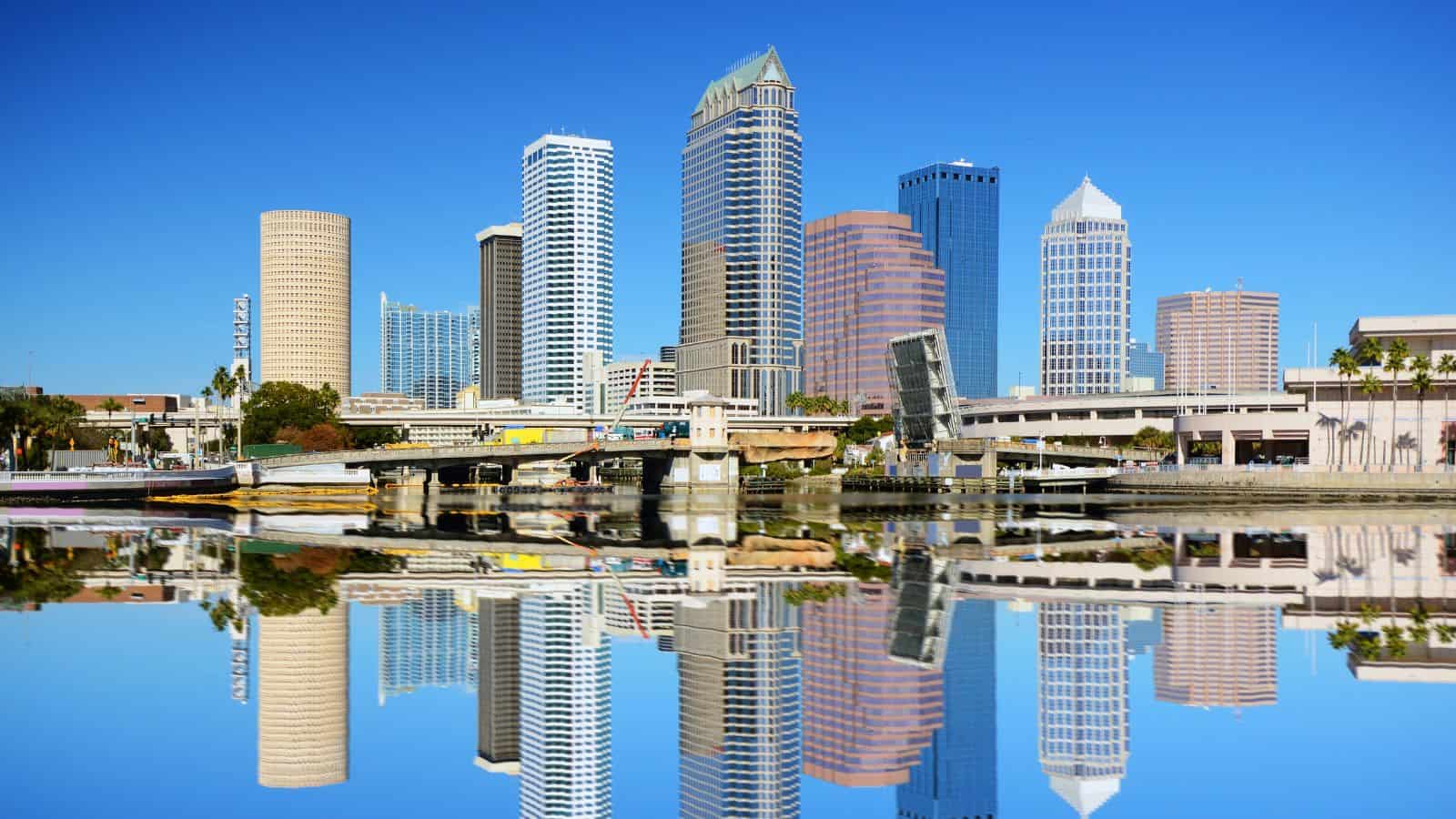 A city skyline with modern high-rise buildings is reflected in calm water under a clear blue sky. There are bridges, palm trees, and a mix of glass and concrete structures along the waterfront.