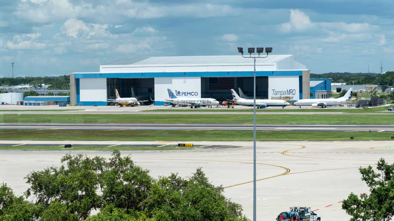 A large aircraft maintenance hangar with the signs “PEMCO” and “Tampa MRO” is seen behind several parked airplanes on the tarmac at an airport under a partly cloudy sky. Trees are visible in the foreground.