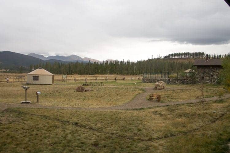 An open field with dry grass, a gravel path, a round yurt, a stone building, large rocks, and distant pine-covered hills under a cloudy sky. A wooden fence runs across the background.