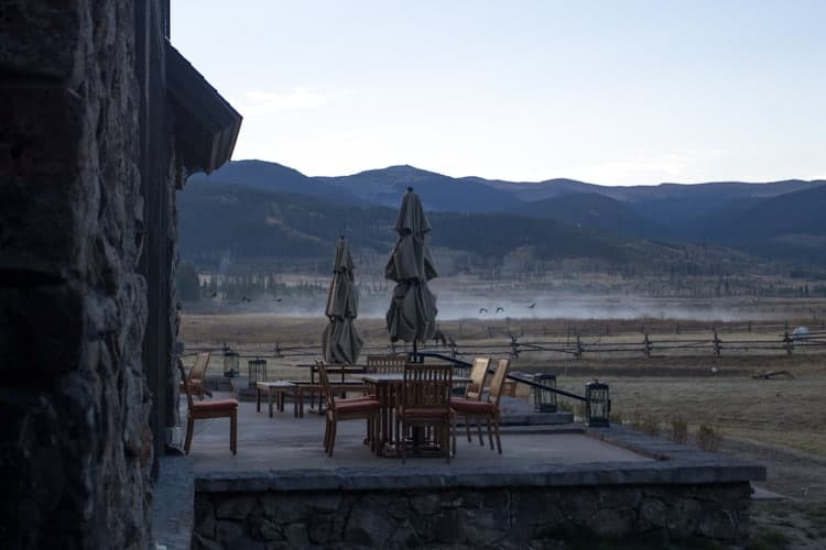 A stone patio with wooden tables, chairs, and closed umbrellas overlooks a misty field and distant mountains under a pale sky at dusk or dawn.