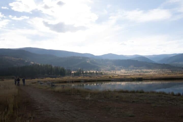 A dirt path runs alongside a calm body of water with grassy fields and trees. Distant mountains rise under a partly cloudy sky. A few people walk on the path to the left.