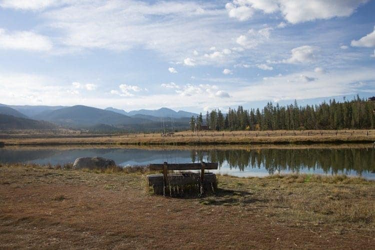 A wooden bench faces a calm pond with a grassy field and trees in the background. Mountains and a partly cloudy sky are visible in the distance. A large rock sits near the water’s edge.