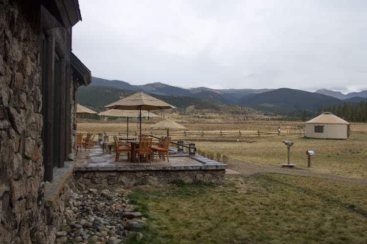A stone building with a wooden patio featuring tables, chairs, and umbrellas overlooks a grassy field with mountains in the background and a yurt off to the right. The sky is overcast.