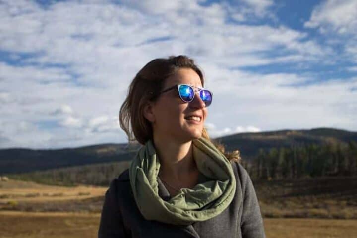 A person wearing blue reflective sunglasses and a scarf stands outdoors in a grassy field with hills and a partly cloudy sky in the background.