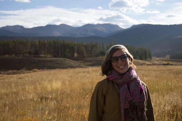 A woman wearing sunglasses, a scarf, and a jacket stands smiling in a dry grassy field with pine trees and mountains in the background under a partly cloudy sky.