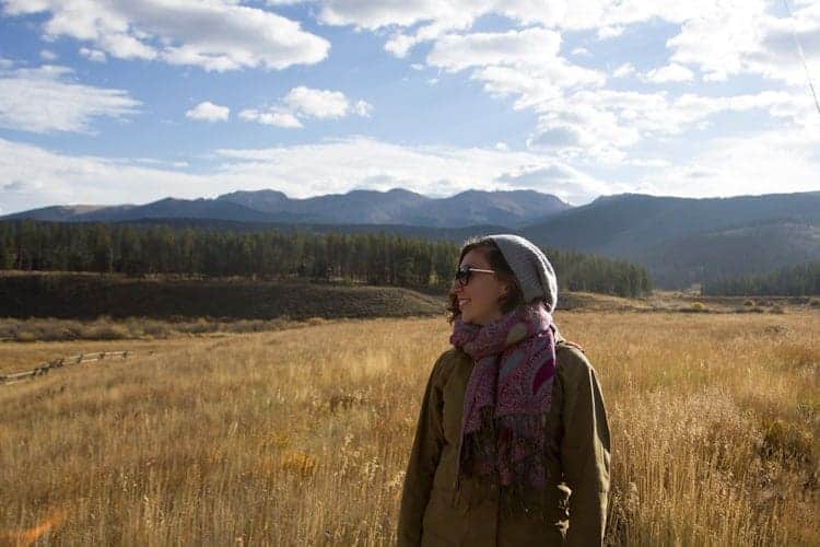 A person wearing sunglasses, a scarf, a beanie, and a jacket stands in a grassy field with mountains and a forest in the background under a partly cloudy sky.