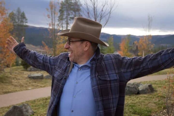 A man wearing a brown cowboy hat, glasses, a blue shirt, and a plaid jacket stands outdoors with arms outstretched, smiling. Autumn trees and distant hills are visible in the background.