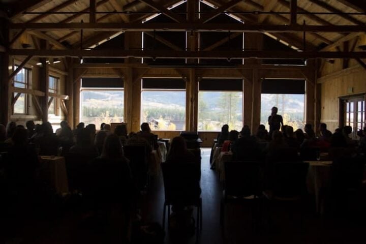 A group of people sits facing a speaker in a large, wooden building with high ceilings and large windows. The outside view shows mountains and trees under a cloudy sky. Light streams in through the windows.
