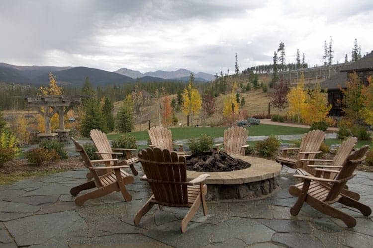 Several wooden Adirondack chairs are arranged in a circle around a stone fire pit on a patio, surrounded by landscaped greenery and trees with mountains visible in the distance under a cloudy sky.
