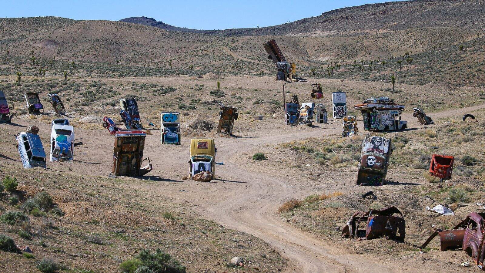 A dirt road winds through a desert landscape where several partially buried, upright cars covered in colorful graffiti are scattered, with dry hills and sparse vegetation in the background.