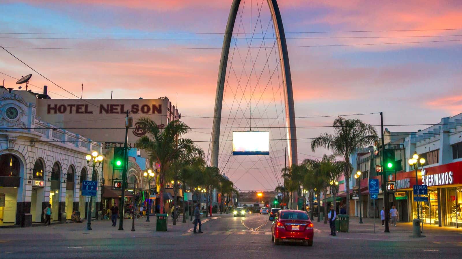 A city street scene at sunset with cars, palm trees, streetlights, and businesses, including Hotel Nelson on the left. A large arch structure is visible in the background under a pink and blue sky.