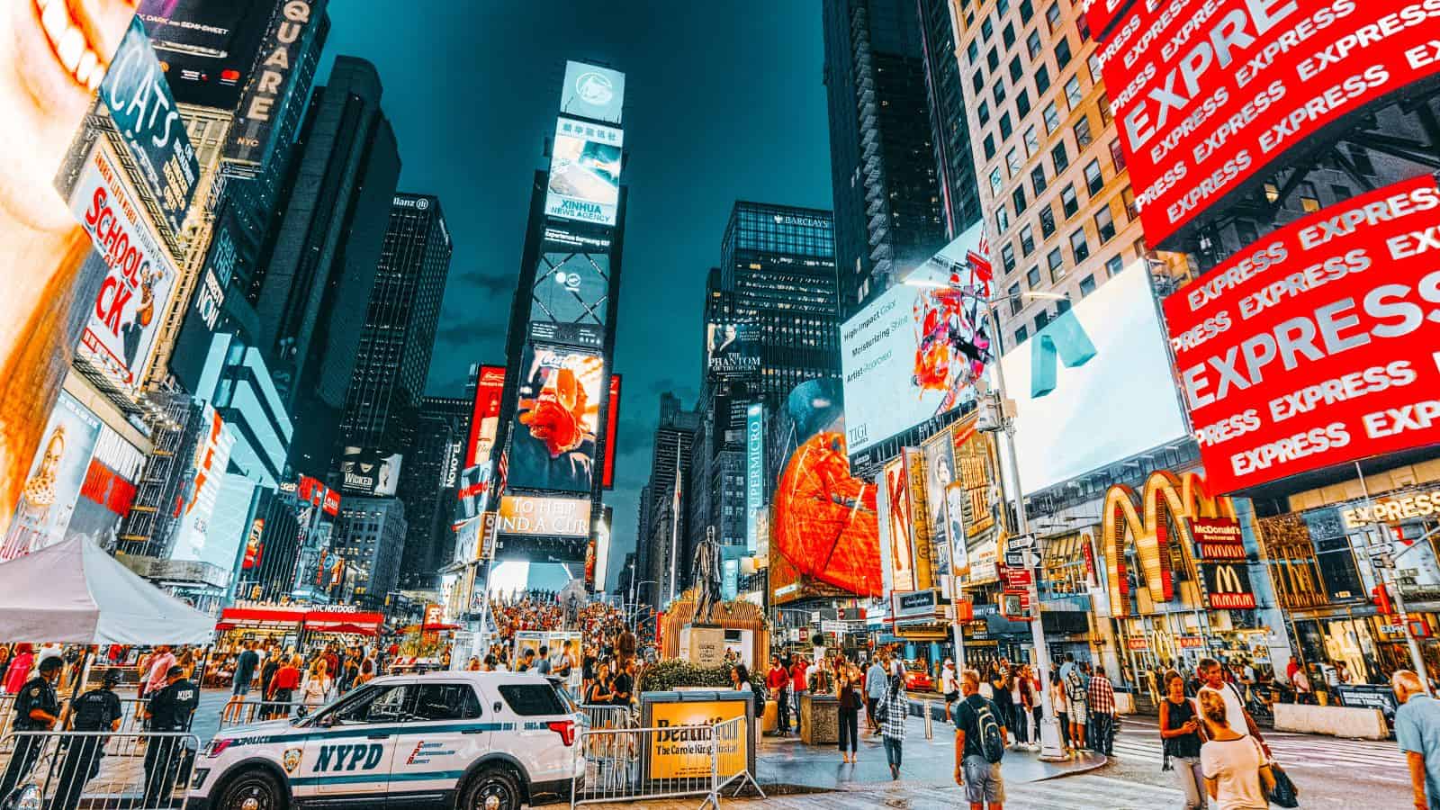A busy Times Square scene at dusk with large electronic billboards, crowds of people, a parked NYPD vehicle, and several recognizable store signs including McDonald’s and Express.