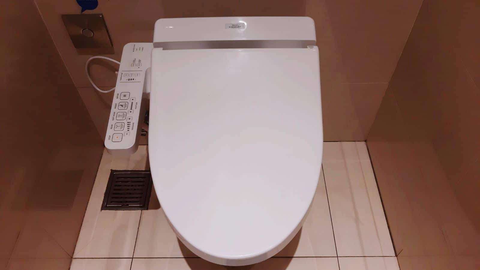 A modern bidet toilet with a control panel on the left side, installed in a tiled bathroom. The toilet seat is closed and the floor has a square drain nearby.