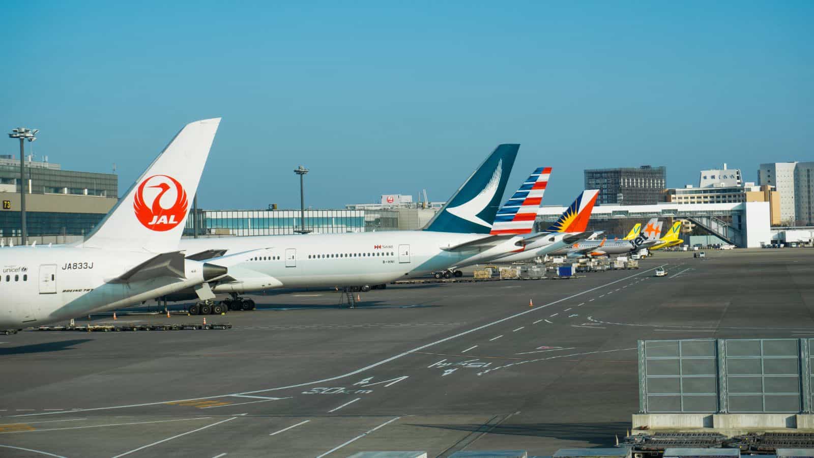 Several commercial airplanes from different airlines are parked at airport gates, with their tails visible and terminal buildings in the background under a clear blue sky.