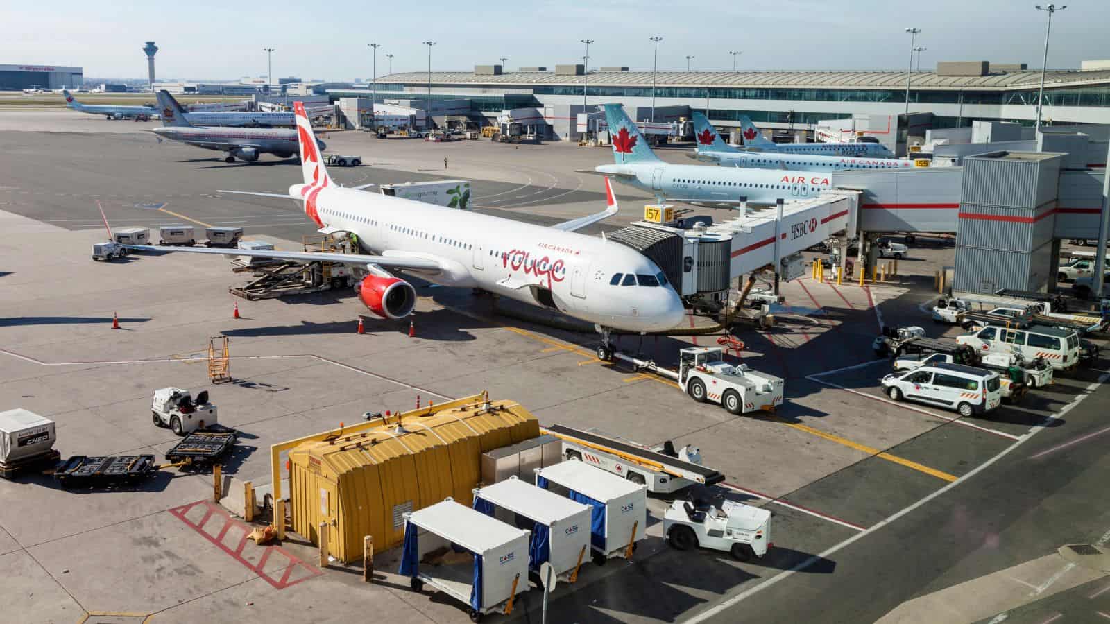 Two Air Canada airplanes are parked at gates at an airport terminal. Ground service vehicles and equipment are positioned around the aircraft. Additional planes and the control tower are visible in the background.