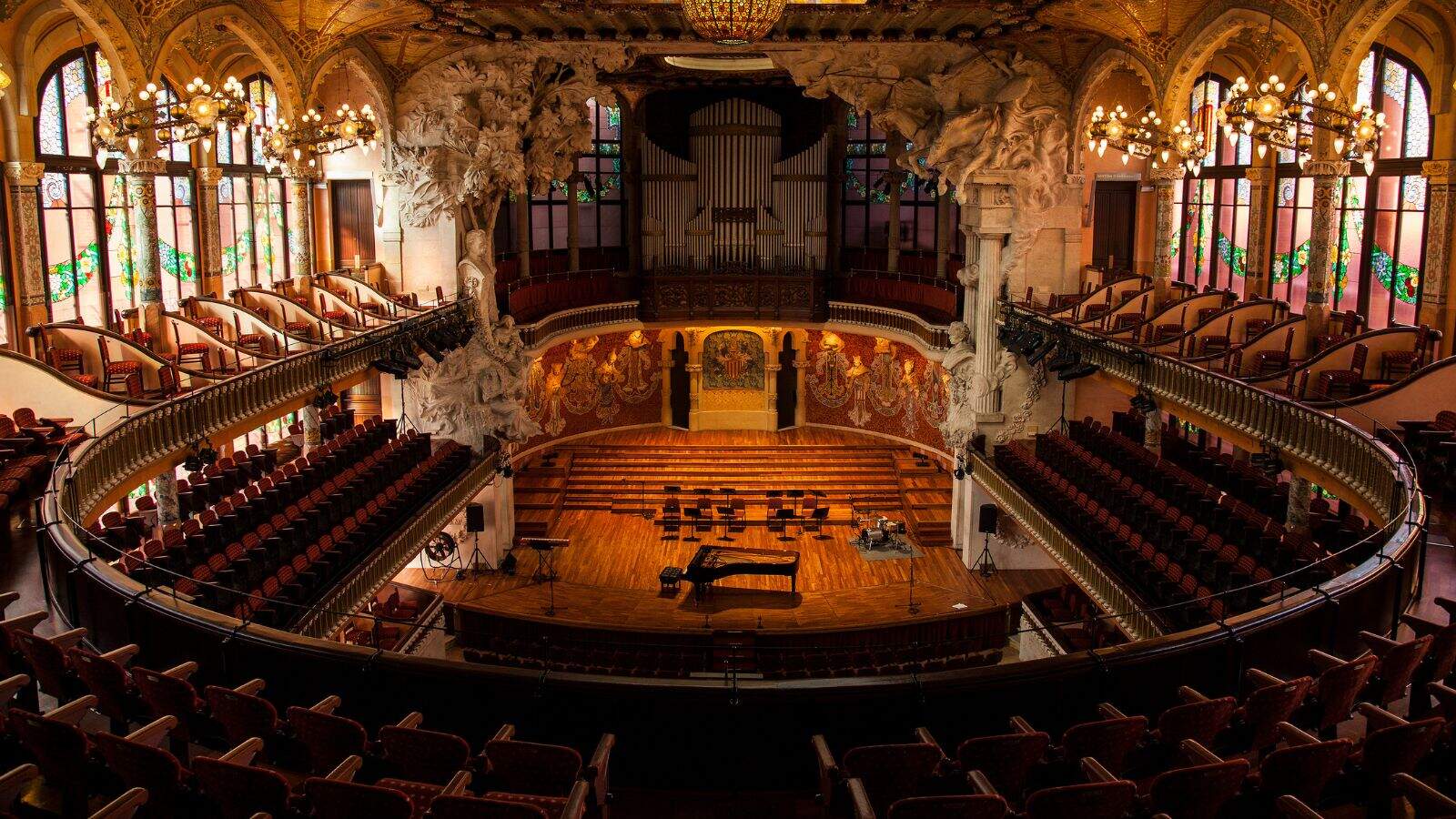 A grand concert hall with ornate architecture, large organ pipes at the back, curved balconies, detailed sculptures, stained glass windows, and a piano on a wooden stage, viewed from the rear of the auditorium.