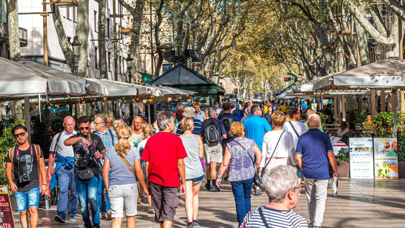 A busy pedestrian street lined with market stalls and outdoor cafes, filled with travelers in Europe walking in both directions under leafy trees on a sunny day.